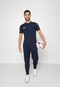 Marine-Ausbildungsausrüstung, bestehend aus einem kurzärmeligen Oberteil mit weißen Akzenten, enganliegender Hose und weißen Fußballschuhen, während ein Fußball gehalten wird.