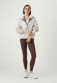 Letvægts sølvjakke med elastiske manchetter og justerbar bund, parret med brune leggins og hvide sneakers. Minimalistisk design.