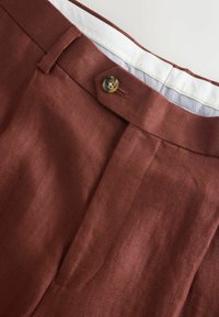 Gros plan sur un pantalon couleur rouille avec une ceinture boutonnée, des passants de ceinture et une doublure intérieure à carreaux partiellement visible.