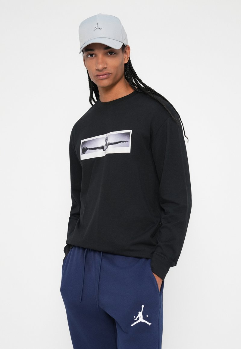 Schwarzes Langarmshirt mit einem großen zentralen Grafikdesign. Das Modell trägt navyblaue Jogginghosen mit einem weißen Jordan-Logo und eine hellgraue Cap.
