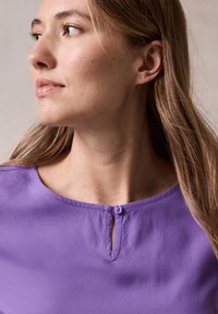 Blouse violette avec un col rond, présentant un petit détail en forme de clé à l'avant, fabriquée en tissu lisse avec une texture douce.