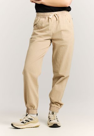 Persoon draagt beige joggingbroek met trekkoord en beige-witte sneakers, staat met gekruiste armen tegen een lichte achtergrond.