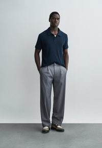 Άνδρας στέκεται με τα χέρια στις τσέπες, φορώντας μπλε σκούρο πόλο πουκάμισο, γκρι πιετέ παντελόνια και δίχρωμα δερμάτινα παπούτσια.