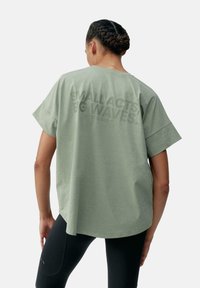 Gaiši zaļš pārāk liels T-krekls ar īsām piedurknēm un apaļu kakla izgriezumu. Aizmugurē drukāts uzraksts "MAZI DARBI, LIELAS VIENĪBAS!" izceltā tekstā.