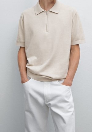 Homme portant un polo beige à manches courtes avec fermeture éclair à l'avant et un pantalon blanc, les mains dans les poches, debout devant un mur clair uni.