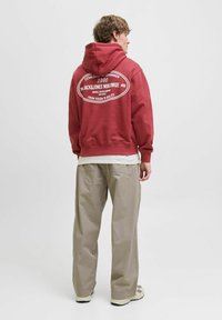 Jeune homme portant un hoodie rouge avec du texte blanc dans le dos, un pantalon beige à jambe large et des baskets blanches, se tenant devant un fond uni.