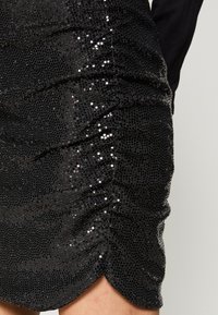 Mini gonna brillante nera con paillettes e dettagli arricciati indossata sulla pelle nuda, abbinata a una maglietta nera a maniche lunghe su uno sfondo neutro.