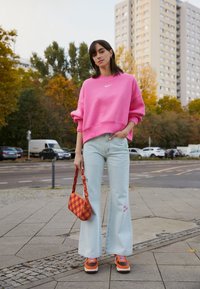 Rosa sweatshirt med logotyp, ljusblå vida jeans med blomsterbroderi, orange och vita sneakers samt en rutmönstrad väska.