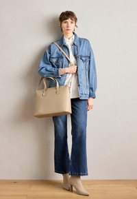 Beige tote bag with dual handles, smooth texture, and minimalistic design. Model wearing a blue denim jacket, white shirt, and flared jeans.