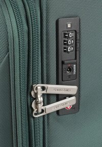 Valigia verde in tessuto con doppie zip in metallo, un lucchetto a combinazione integrato e una caratteristica di lucchetto approvato TSA. Superficie testurizzata con linee sottili.