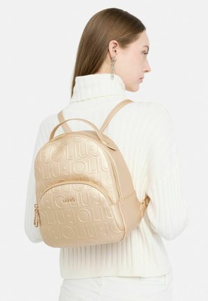 Zaino in pelle beige compatto con logo impresso, forma arrotondata, spallacci regolabili e tasca con zip. Superficie testurizzata con dettagli lisci.