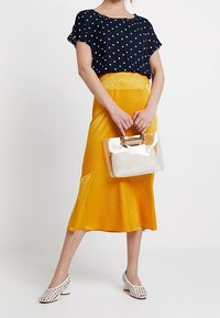Námořnický top s puntíky, žlutá saténová midi sukně, bílé boty s výřezem a průhledná kabelka s bílým akcentem. Hladké textury a výrazné barvy.