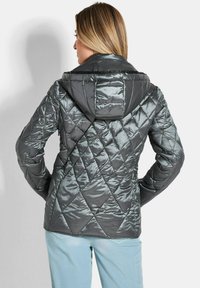 Dunkelgraue gepolsterte Jacke mit glänzender Oberfläche. Verfügt über ein Rautenmuster und eine Kapuze. Getragen mit hellblauen Hosen.