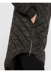 Giacca imbottita verde scuro con orlo asimmetrico, zip laterale e polsini a coste. Il tessuto appare lucido con una texture liscia e un motivo a diamante.