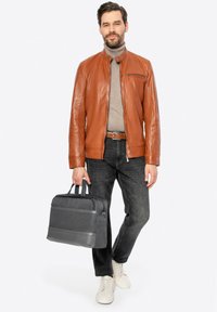 Giacca di pelle marrone con leggera lucentezza, camicia grigia a maniche lunghe, jeans in denim scuro e borsa grigia testurizzata. Sneakers bianche completano l'outfit.