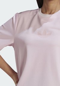 Žena ve světle růžovém tričku Adidas s tónovým logem Trefoil, pózující s jednou rukou na boku před jednobarevným pozadím.
