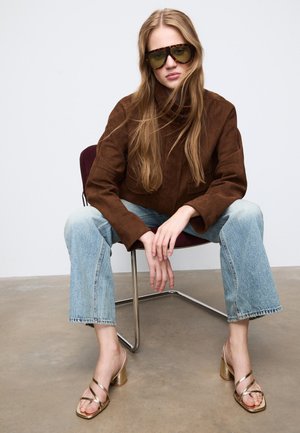 Hnědá semišová bunda, světle modré zvonové džíny a zlaté sandály na blokovém podpatku. Převelké zelené sluneční brýle dokončují outfit.