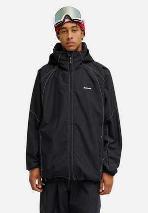Junger Mann, der eine schwarze Schneejacke mit weißen Nähten, eine rote Mütze und Skibrille auf der Stirn trägt, steht vor einem schlichten Hintergrund.