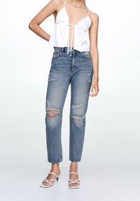 Bílý crop top s krajkovým lemem a šňůrkou, spárovaný s světle modrými vysoce pasovými džíny s roztrženinami. Na nohou bílé sandály na pásky.