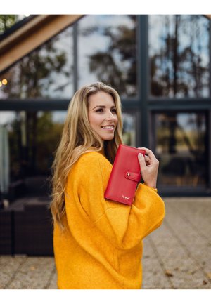 Red leather wallet with a snap closure, holding in right hand, set against a backdrop of large windows and wooden architecture.