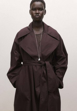 Modelo vistiendo un abrigo trench oscuro oversized con cinturón y un collar largo con un colgante de plata, posando contra un fondo liso claro.