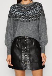 Femme portant un pull en tricot gris à motifs avec des manches longues et une mini-jupe en simili cuir noire à boutons.