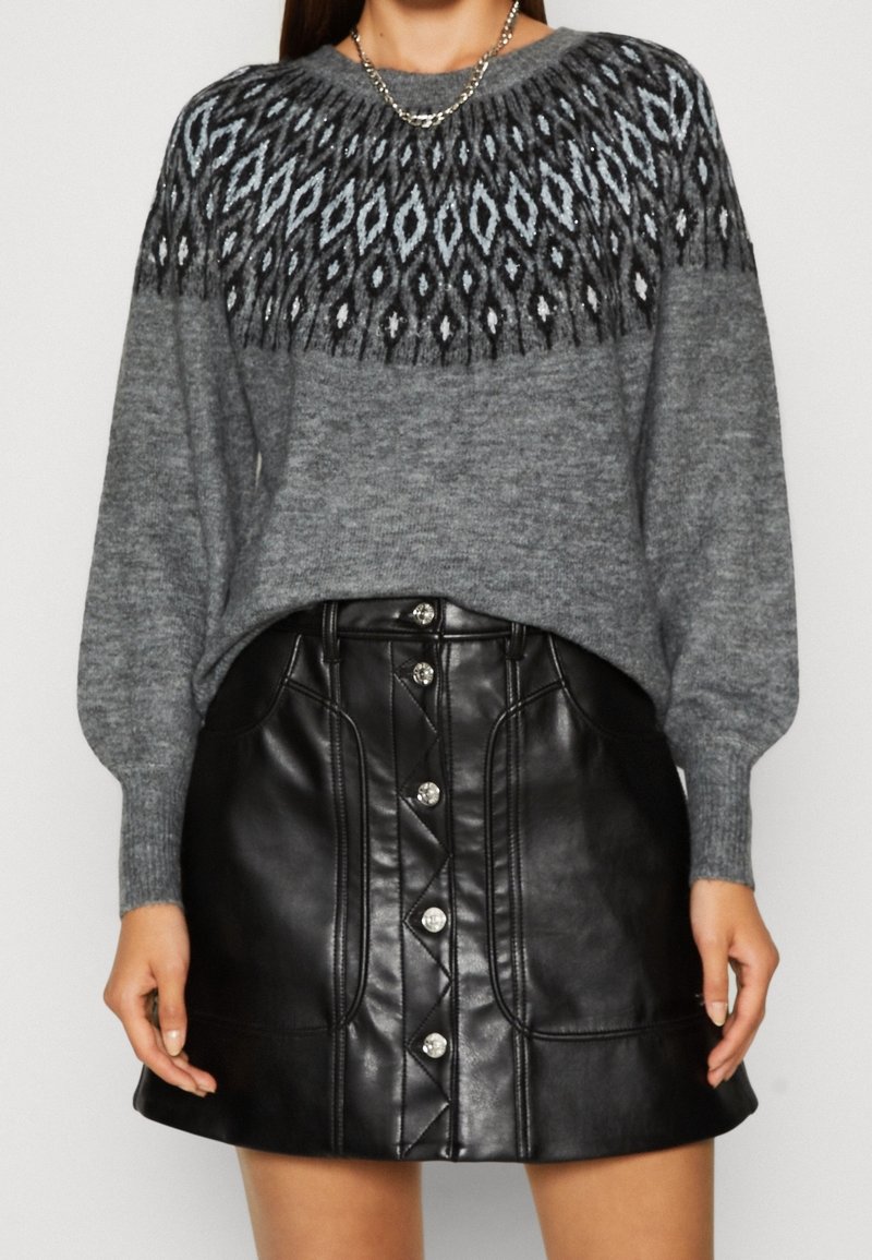 Femme portant un pull en tricot gris à motifs avec des manches longues et une mini-jupe en simili cuir noire à boutons.