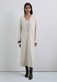 Vestido de punto beige con escote en V, mangas largas y puños acanalados, que presenta una textura sutil y aberturas laterales. Combinado con botas marrones hasta la rodilla.