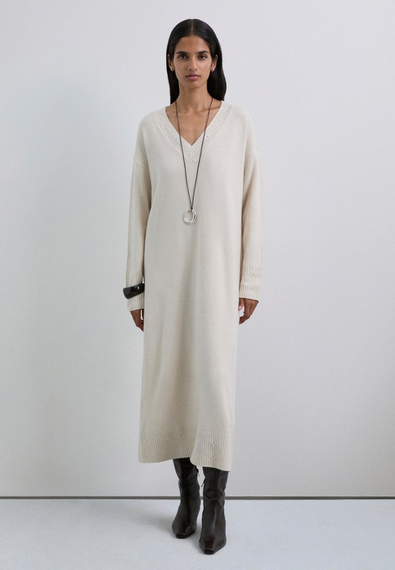 Vestido de punto beige con escote en V, mangas largas y puños acanalados, que presenta una textura sutil y aberturas laterales. Combinado con botas marrones hasta la rodilla.