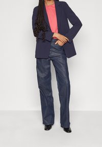 Kvinna klädd i en marinblå kavaj, korallrosa skjorta, vida mörk jeans och svarta stövlar, stående mot en neutral bakgrund.