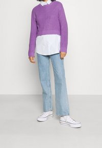 Fioletowy prążkowany krótki sweter noszony na białej koszuli zapinanej na guziki, zestawiony z jasnoniebieskimi rozszerzanymi dżinsami i białymi trampkami.