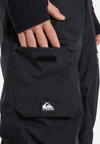 Hand die in een zwarte cargozak met klittenbandsluiting van zwarte broek steekt, met daarop een Quiksilver-logo patch.