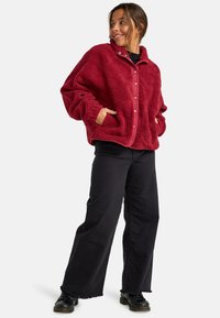 Giacca in pile rossa con colletto alto, apertura con bottoni a pressione e due tasche laterali, abbinata a pantaloni neri a gamba larga e scarpe nere.