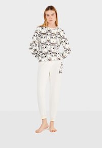 Conjunto de pijama blanco que incluye una camiseta de manga larga con un patrón de animales de dibujos animados repetido y pantalones blancos lisos con un detalle gráfico lateral.
