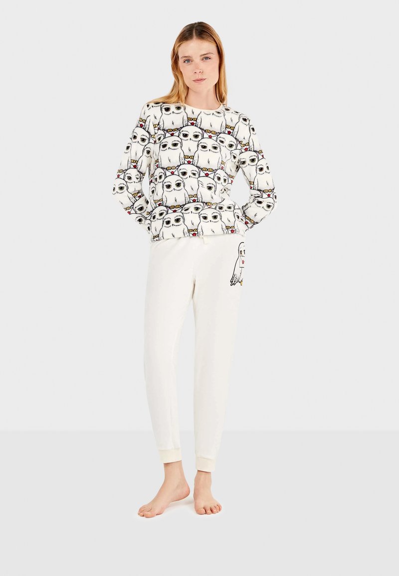 Conjunto de pijama blanco que incluye una camiseta de manga larga con un patrón de animales de dibujos animados repetido y pantalones blancos lisos con un detalle gráfico lateral.