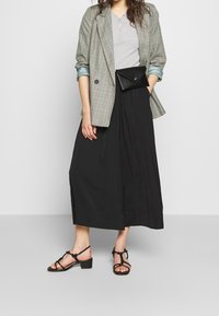 Blazer a cuadros en verde y gris, camiseta gris, falda plisada negra y sandalias de tacón con tiras negras. Un bolso de mano negro complementa el atuendo.