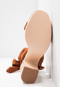 Brun ruskind højhælet sko med en struktureret korkhæl og en glat gummisål, vippet mod en hvid væg. Detaljerne inkluderer en sløjfedetail.