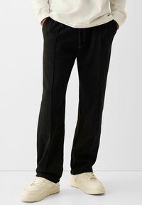 Black sweatpants with an elastic waistband, side pockets, and a straight-leg design, paired with beige sneakers. Smooth texture.