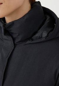 Czarna kurtka puchowa z wysokim kołnierzem, gładką fakturą i detalem w postaci guzików z przodu. Materiał wygląda na ocieplany i elegancki.