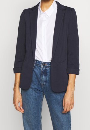 Kobieta ubrana w białą koszulę na guziki, granatową teksturowaną marynarkę z podwiniętymi rękawami oraz niebieskie jeansy z wysokim stanem na jednolitym tle.