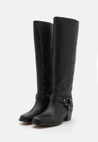 Bottes noires en cuir montantes jusqu'au genou, à texture lisse, avec une lanière latérale ornée d'accents métalliques et un talon épais.