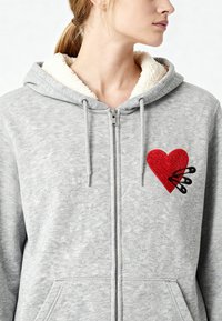 Felpa grigia con zip e cappuccio foderato in Sherpa. Presenta una toppa a forma di cuore rosso con spilloni neri. Tessuto testurizzato e tasche frontali incluse.