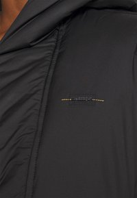 Černá bunda s hladkou, matnou texturou, výrazným prošíváním a nášivkou s logem a žlutými akcenty na kapsičce na hrudi.