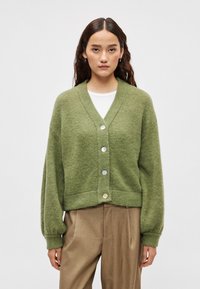 Πράσινη πλεκτή ζακέτα με V-λαιμόκοψη, πέντε κουμπιά και ριμπ μανσέτες. Η υφή είναι απαλή, ελαφρώς χνουδωτή και κομμένη στο ύψος της μέσης.