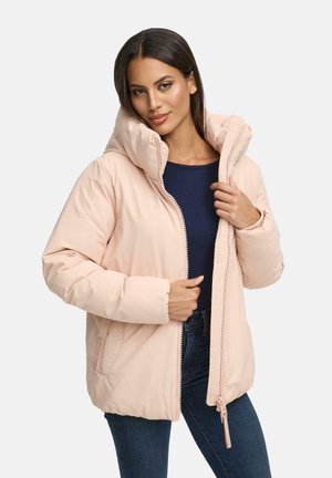 Vrouw in een lichtroze donsjack over een marineblauwe top en donkere spijkerbroek, die de rits van het jack gedeeltelijk gesloten houdt.