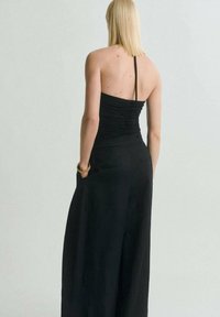 Jumpsuit noir sans manches avec un corsage ajusté et des jambes larges, comportant des détails froncés à l'arrière et des poches latérales. Texture lisse.