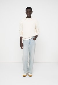 Young man wearing a cream knit sweater, light blue jeans, and white sneakers, standing with one hand in pocket against white background.