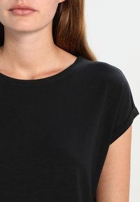Žena s bledou pletí a dlouhými hnědými vlasy, oblečená v jednoduchém černém tričku s krátkým rukávem a kulatým výstřihem.