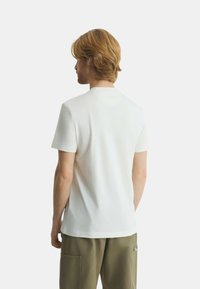 T-shirt bianca a maniche corte con collo a giro, texture liscia, design aderente e orlo dritto, indossata con pantaloni verdi oliva.