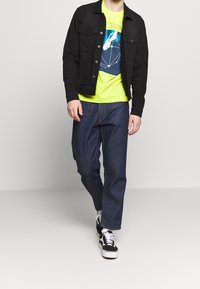 Černá džínsová bunda, neonově zelené tričko s grafikou a tmavě modré široké džíny. Černé tenisky s bílými tkaničkami dokončují outfit.